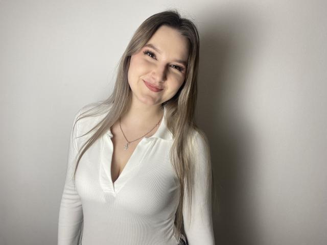 Karpinka with long blonde hair, smiling, wearing a white, v-neck, long-sleeve shirt, showing small tits, standing against a plain wall. She's cute, with a subtle, sexy look.