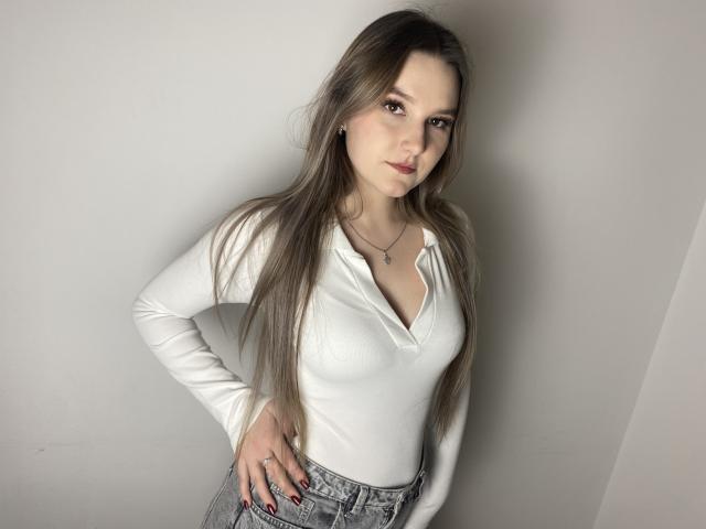 Karpinka with long brown hair, white long-sleeve top, showing small tits, hand on hip, gray jeans, necklace, and red nail polish, standing against a plain wall.