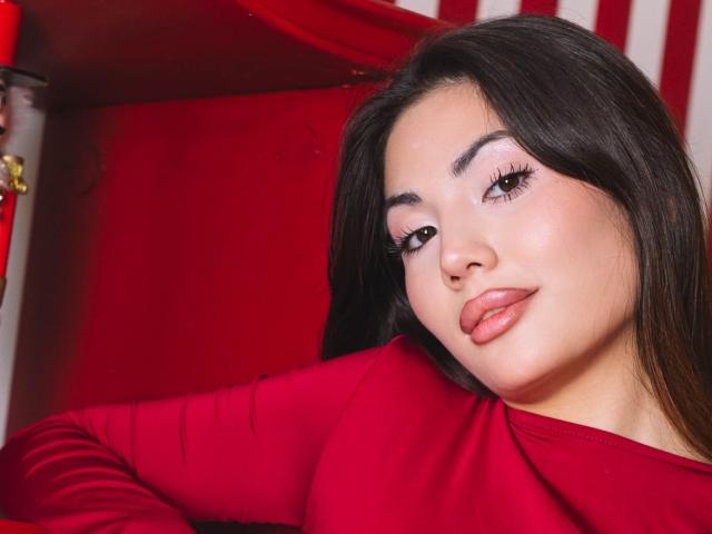 MonicaLuxe mit dunklem Haar, roten Lippen und langen Wimpern, trägt ein rotes Oberteil und zieht leicht die Lippen zusammen, vor einem rot-weiß gestreiften Hintergrund. Die Brüste sind teilweise sichtbar..