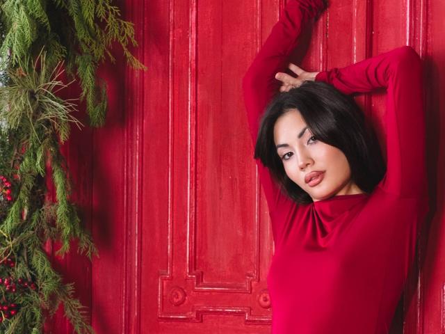 MonicaLuxe, avec des cheveux foncés, un haut rouge à manches longues, les bras levés, regardant la caméra, une porte en bois peinte en rouge, un sapin de Noël avec des branches vertes et des baies rouges sur la gauche..