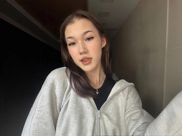LixDomii with brown hair, light skin, and subtle makeup, wearing a gray hoodie, black shirt, and small necklace, has a slight pout and a small beauty mark on her cheek.