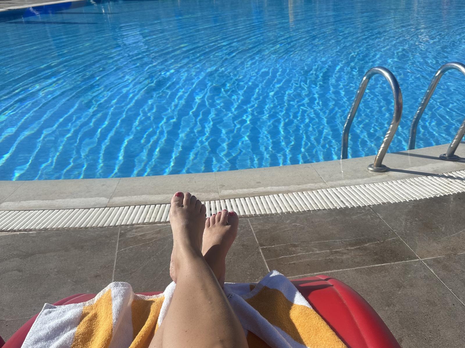 Ballerina69's feet with red-painted toenails, crossed at the ankles, resting on a red pool chair with a yellow towel, looking over a clear blue swimming pool.