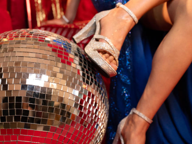 VanesaValet's sparkly silver high heels with ankle straps rest on a shiny, reflective, red-and-blue mosaic ball, her tan legs in a blue sequined dress.