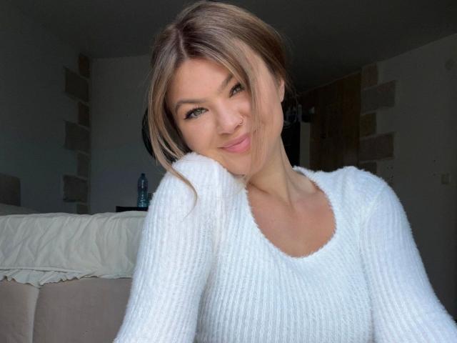 Avaexklusiv-hot with blonde hair and green eyes, wearing a white, fluffy, ribbed sweater, smiling softly, head resting on her right hand.
