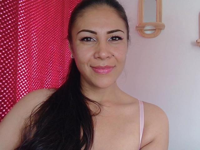 KimmyHolleshot with dark hair in a ponytail, pink lipstick, and brown eyes, smiling at the camera, wearing a pink spaghetti strap top, showing her tits.