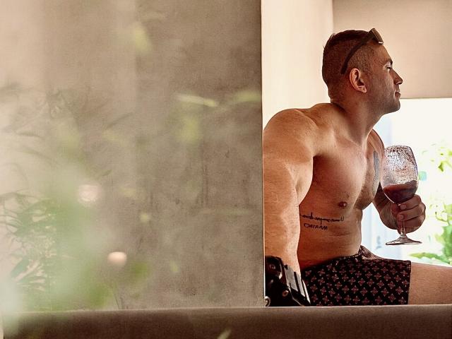 PaulHotS, shirtless, muscular, holding wine glass, star-patterned shorts, tattoo on right side, brown hair, looking outside, slight smile, standing by concrete wall, daytime.