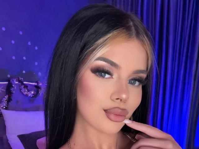 IvyRaven has black hair with blonde highlights, blue eyes, thick eyebrows, and pink lipstick. She's touching her lips with her finger, looking at the camera.