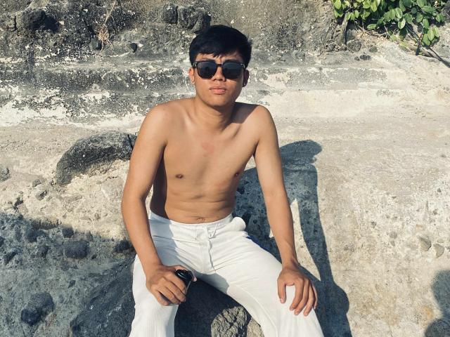 Shirtless Rayzen69 with tan skin, black hair, and sunglasses, wearing white pants, sitting on a rock, holding a black watch, with a slight tan line on his chest.