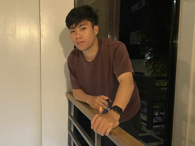 Rayzen69, leaning on a railing, wearing a brown shirt and black wristwatch, with short black hair, and a slightly serious expression, in a dimly lit room.