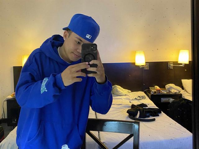 Rayzen69 wearing a blue hoodie and matching blue cap, taking a mirror selfie with a camera, smiling slightly, standing in a messy bedroom with a bed and lamps.