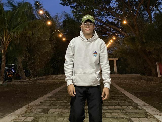 Rayzen69 with glasses, green cap, white hoodie, black pants, standing on cobblestone path, string lights overhead, trees and palm in background, dusk sky, neutral expression.