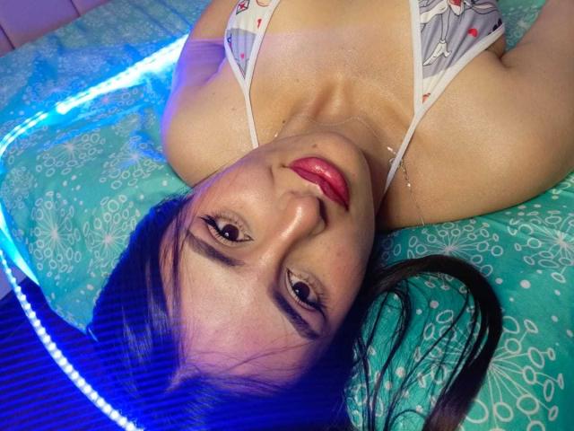 BrendalX is lying on a bed with blue LED lights, head upside down, looking at the camera. She has red lipstick, black hair, and is wearing a white, patterned top.