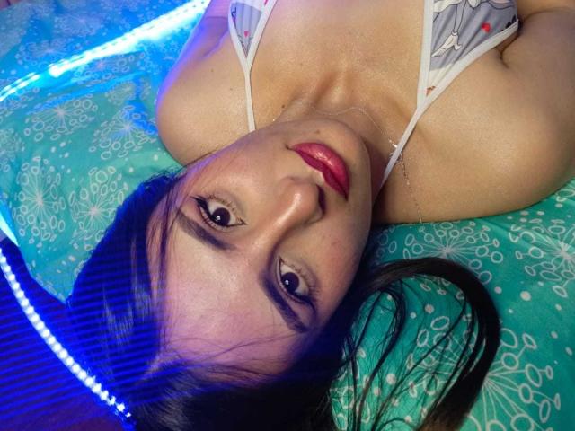 BrendalX's face is upside down, looking at the camera with red lipstick, brown eyes, and black hair, wearing a white bra with gray and red patterns, on a blue bed.