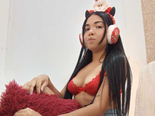 AliceSmity with long black hair, wearing red lace bra and Christmas headphones with Santa ears, holding red fuzzy pillow, has small tits, cute and confident, sitting down.