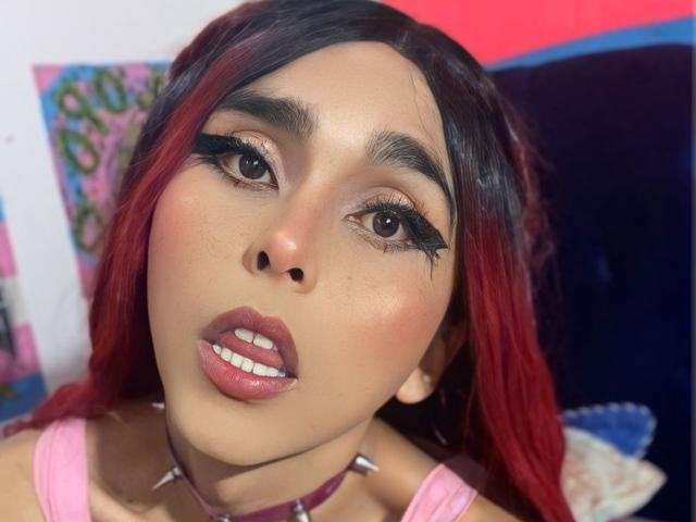 Zasha69 with red and black hair, dark eyeliner, and pink lipstick, wearing a pink tank top and purple choker, looking directly at the camera with slightly parted lips.