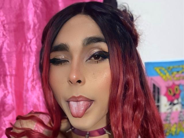 Zasha69 with red and black ombre hair, winking brown eyes, and tongue out, wearing a pink choker, has a cute face and slightly shiny skin.