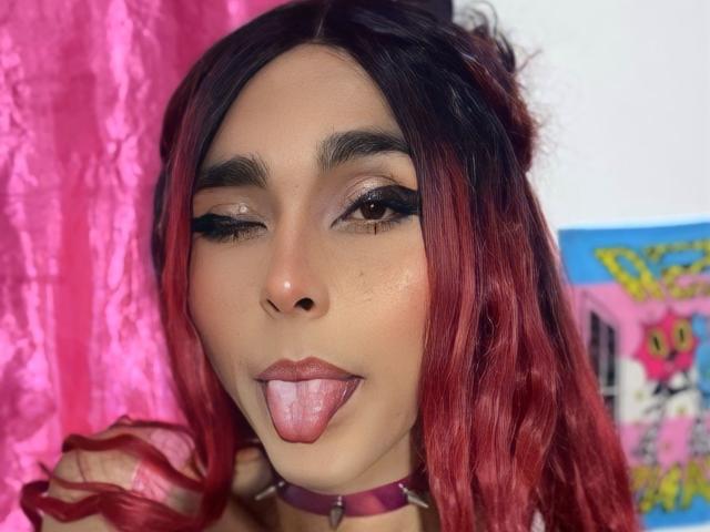 Zasha69 with red and black hair, winking, sticking out tongue, dark eyeliner, choker, pink curtain, and shiny skin, showing off her cute face.