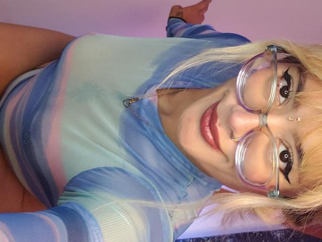 FreankyQueen with blonde hair, blue tie-dye top, and glasses, smiling at camera, nose ring, and forehead dot, showing her tits slightly. Her ass is barely visible.