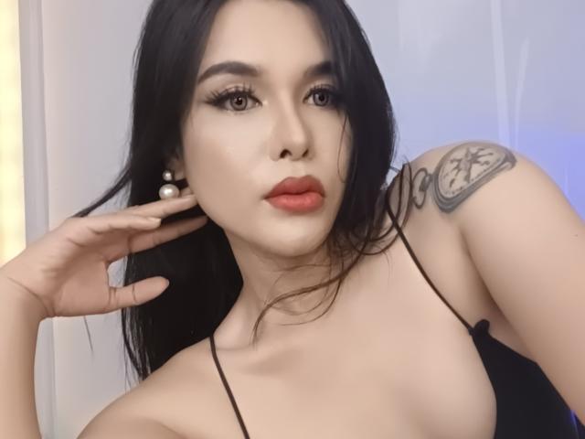 MyLuvNavi has long black hair, red lipstick, and a tattoo on her right shoulder. She's wearing a black spaghetti-strap top, showing her tits, and pearl earrings.