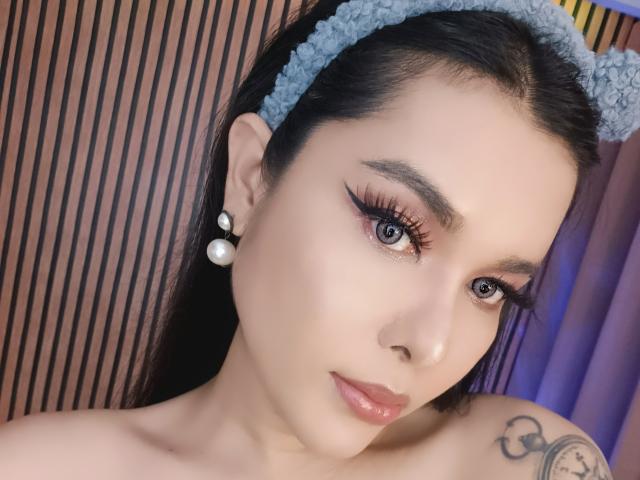 MyLuvNavi has pale skin, blue eyes, and long black hair with a gray headband. She's wearing pearl earrings, has winged eyeliner, and has glossy pink lips. Tattoo on right shoulder.