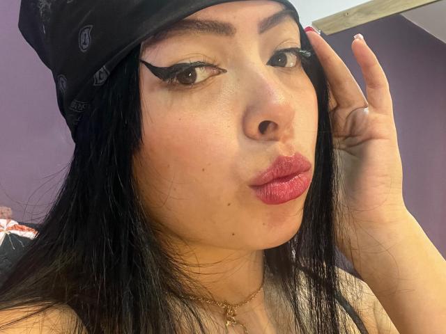 KateWelsin with black hair, wearing a black bandana, thick black eyeliner, red lipstick, touching her head, gold necklace, purple walls, and a slightly shiny face.