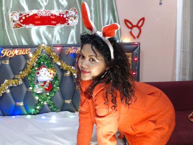 FelyLove met krullend zwart haar, een oranje outfit en witte konijnenoren, knielend op een bed, lachend naar de camera, waarbij haar schattige achterkant te zien is. Kerstdecoratie en de Kerstman op de achtergrond..