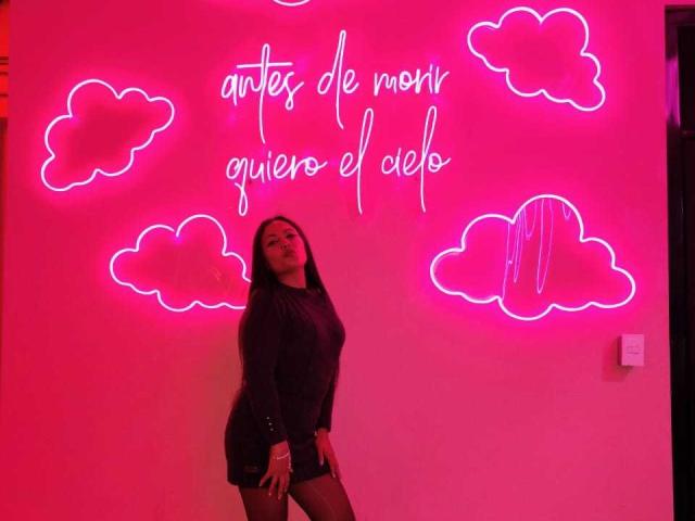CarolineVoss stands with a pink neon wall, wearing a dark long-sleeve dress, black shorts, and a confident smile. Neon clouds and pink lights surround her.