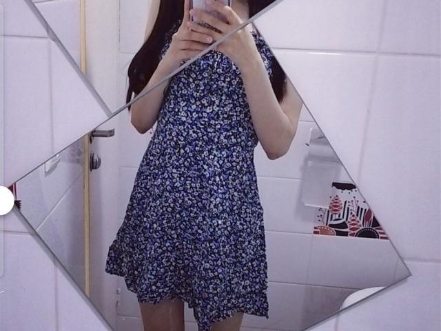 EvaRouseX wearing a blue floral dress, standing in front of a mirror, holding a phone, with visible arms and lower face, but no visible boobs or ass.