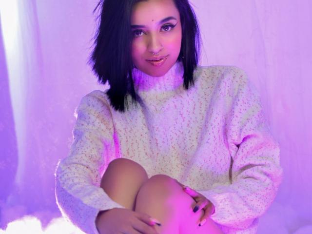AtheneaOwenss with shoulder-length black hair, white knitted sweater, and nose ring, sitting with knees up, purple lighting, smiling subtly, hands on knees, pink nose stud.