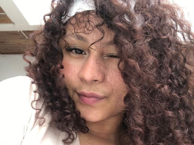SofiaPassionne with curly brown hair, white headband, winking, and slightly puckered lips, wearing a white shirt, in a selfie. Her nose is small and her skin is light.