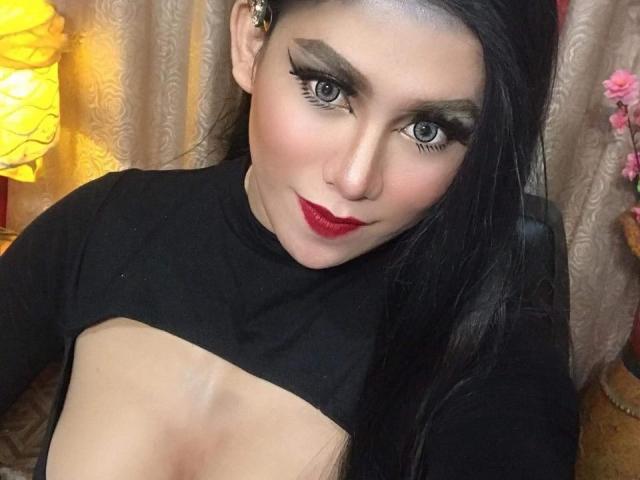 SeductiveGab with dark hair, heavy eyeliner, red lipstick, black top exposing her bare tits, slightly smiling, in a dimly lit room, yellow lamp on the left.