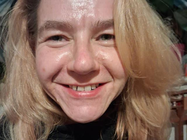 BebeHt's smiling face with blonde wavy hair, slightly shiny skin, and visible wrinkles, showing teeth in a natural, relaxed look. BebeHt's nose is centered with small, brown eyebrows.