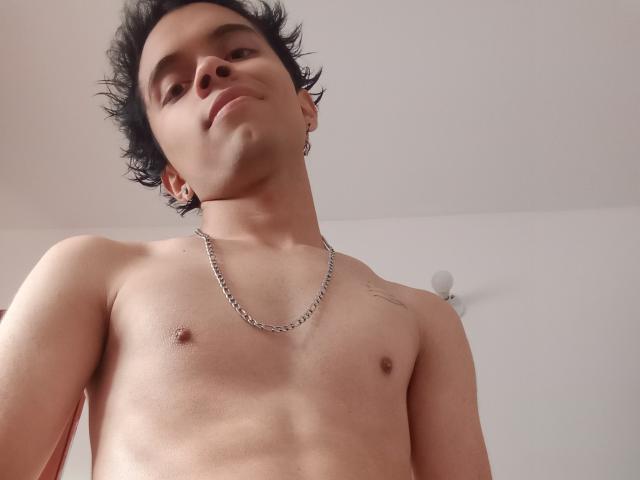 MinhoReyes, shirtless, with short black curly hair, wearing a chain necklace, has a small tattoo on the right shoulder, and is looking down with a slight smirk.