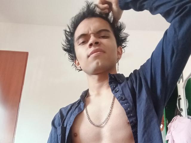 MinhoReyes with spiky black hair, wearing an unbuttoned blue shirt, chain necklace, and ear gauges, looking smug, raising right arm. Naked chest visible, standing in a room.