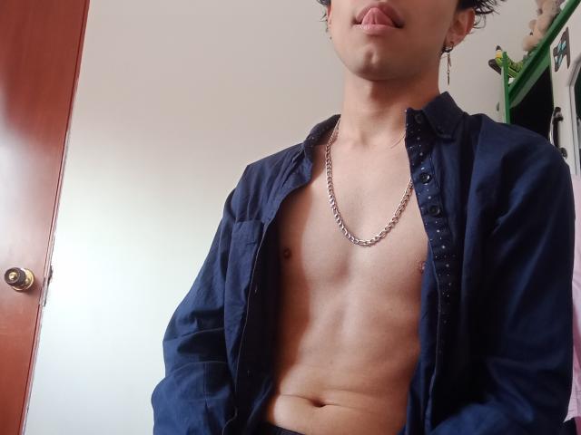 MinhoReyes, shirtless, wearing a blue button-up, chain necklace, sticking out tongue, small nipples, slightly chubby, brown skin, short curly hair, standing in front of a wooden door.