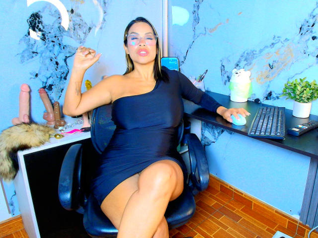 AlixLow, wearing a black one-shoulder dress, sits in a chair with one leg crossed, holding a cigarette. Her glasses reflect the camera. A dildo and keyboard are nearby.