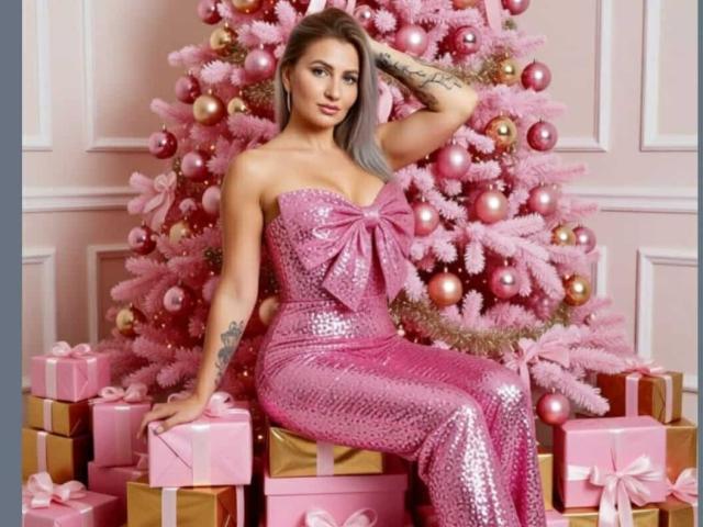ChaudeEvely sits in front of a pink Christmas tree, wearing a strapless, pink sequined dress with a large bow. Her right hand rests on her head, showing a tattoo on her forearm. The tree is decorated with pink and gold ornaments. Several pink and gold wrapped gifts are stacked around her. ChaudeEvely has long, light brown hair with gray streaks, and her makeup is bold with pink lipstick. The overall composition is festive and glamorous.