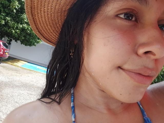 Close-up of MarilynHills, a young woman with wet black hair and a brown straw hat. She has light brown skin with small acne blemishes and a slight smile. She wears a blue tie-dye bikini top. Her right ear has a small hoop earring. The background shows a pool and a red car. The image has a casual, sunlit feel. Her wet hair suggests she's just come from swimming. The close crop emphasizes her facial features and upper torso.