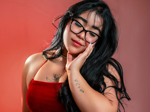 DayannaCold mit langem, schwarzem Haar, trägt ein rotes Kleid, eine schwarze Brille, berührt ihr Gesicht, hat ein kleines Tattoo am rechten Handgelenk, ein leichtes Lächeln, roter Hintergrund, weiches Licht, selbstbewusster Ausdruck..