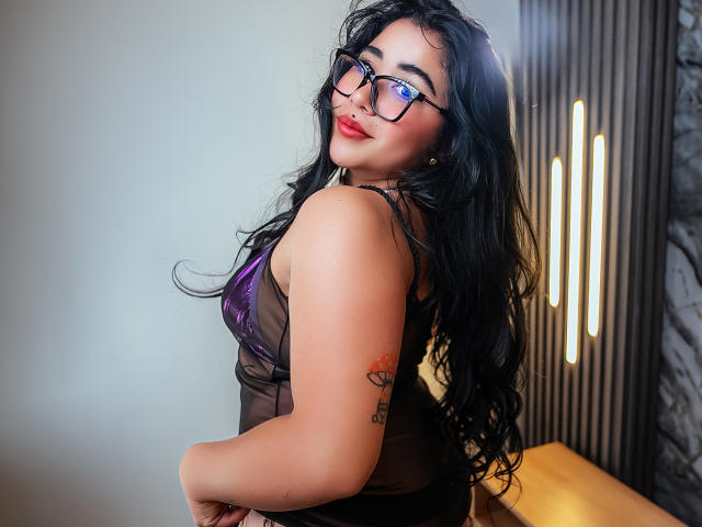 DayannaCold with long black hair, glasses, and red lipstick, wearing a sheer black top, showing her tits and a tattoo on her upper arm. She's looking over her shoulder.