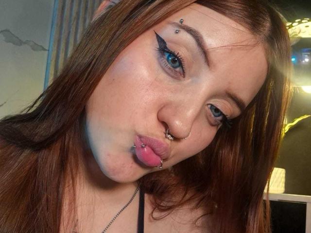 MaylinSanchez with brown hair, blue eyes, and nose piercings, pouting with a tongue piercing, wearing black eyeliner and a nose ring, looking into the camera.