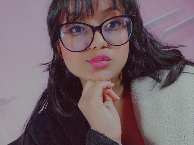 AzulMoon with dark, wavy hair, wearing black glasses, pink lipstick, black jacket with white furry collar, resting chin on hand, looking at camera, soft pink background.