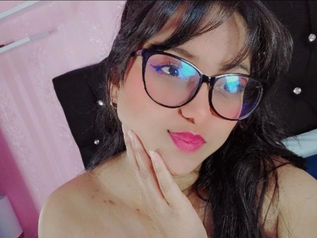 AzulMoon mit dunklem Haar, die eine schwarze Brille und pinken Lippenstift trägt, mit leicht gerötetem Gesicht, die sich an ihr Kinn fasst und eine kleine Prellung an ihrer Schulter zeigt..