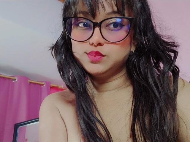 AzulMoon, con cabello negro largo, gafas y lápiz labial rosa, mirando a la cámara con un ligero puchero, mostrando sus hombros desnudos, con cortinas rosas de fondo..