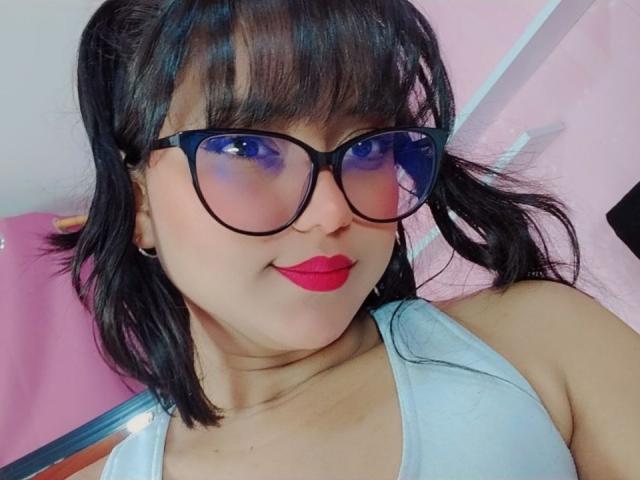 AzulMoon with black pigtails, blue-tinted glasses, red lipstick, and a light grey tank top showing her boobs. She's smiling subtly, with black ear holes in her ears.