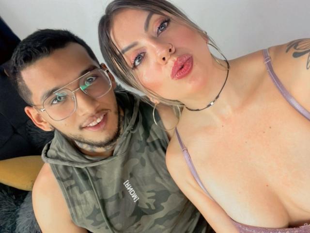 ArianaWil with big tits, pink lipstick, and a choker, pouting, next to a cute, nerdy dude in glasses and a camo hoodie, smiling. Both have light skin.