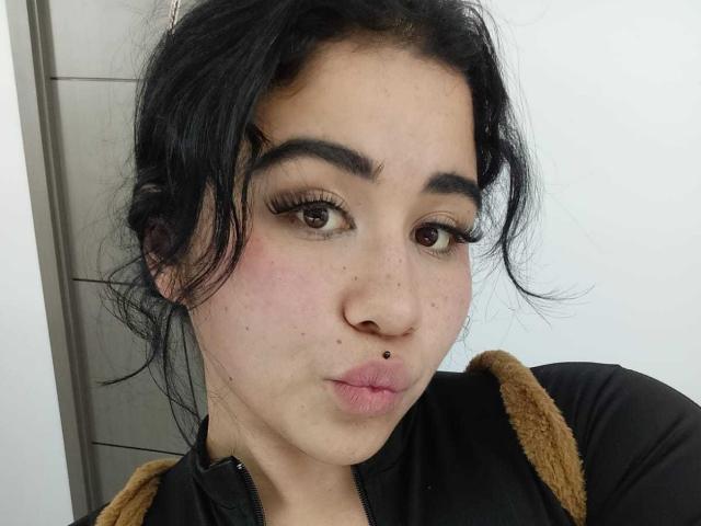 DayannaCold has black hair, brown eyes, and a small nose piercing. She's pouting with long eyelashes, wearing a black top with brown straps. Her face is slightly flushed.