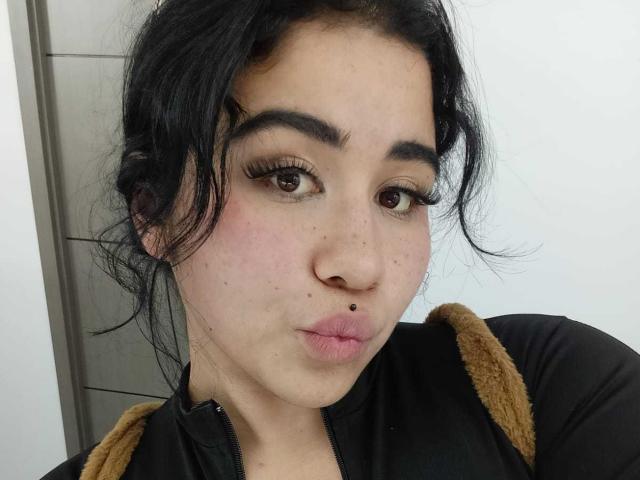 DayannaCold has dark hair, brown eyes, and a small nose boogey. She's pouting with her lips slightly up, wearing a black shirt with brown shoulder straps.