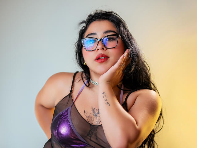 DayannaCold with black hair, wearing purple shiny lingerie, black mesh, glasses with blue reflective lenses, hand on cheek, tattoo on wrist, and gold earrings, looking at camera.