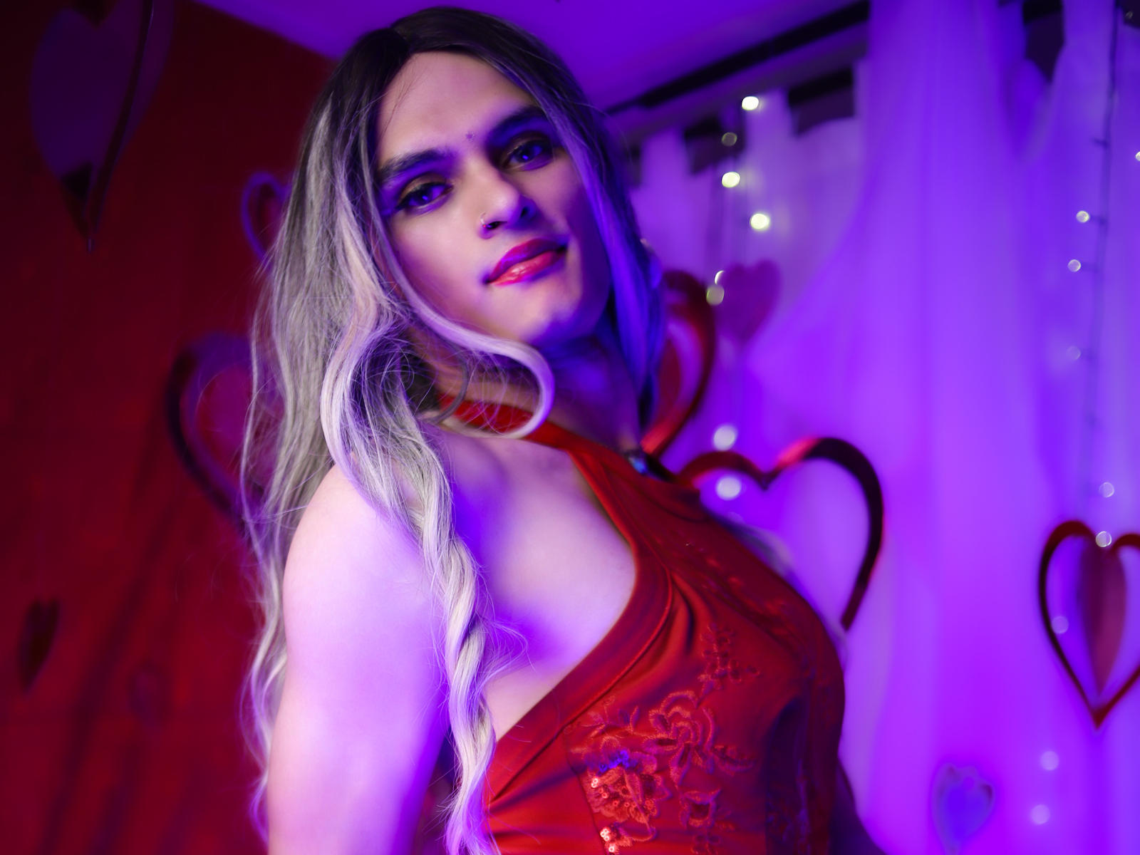 Sabrina Turneer, con lunghi capelli biondi, indossa un abito rosso in pizzo con spalline, con luci viola e blu, sorride alla fotocamera, mostrando un corpo snello e tonico e un viso carino..
